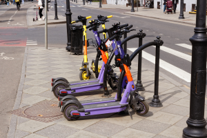 Eine Reihe von Elektro-Scootern, die entlang einer Straße mit Fußgängern, Radfahrern, einem Müllcontainer im Vordergrund und Gebäuden, Bäumen und Pflanzen im Hintergrund geparkt sind.