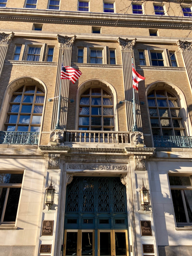 Außensicht des Plaza Hotels in New York City, mit Glasfenstern, Türen, Säulen, Geländern, Leuchten, Texttafeln und Flaggen vor einem Himmelhintergrund.