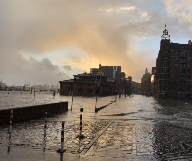 Überflutete Straße in Hamburg, Deutschland mit Wasser auf der Straße, Pfählen, Schildern, Gebäuden und einer Brücke unter einem bewölkten Himmel.
