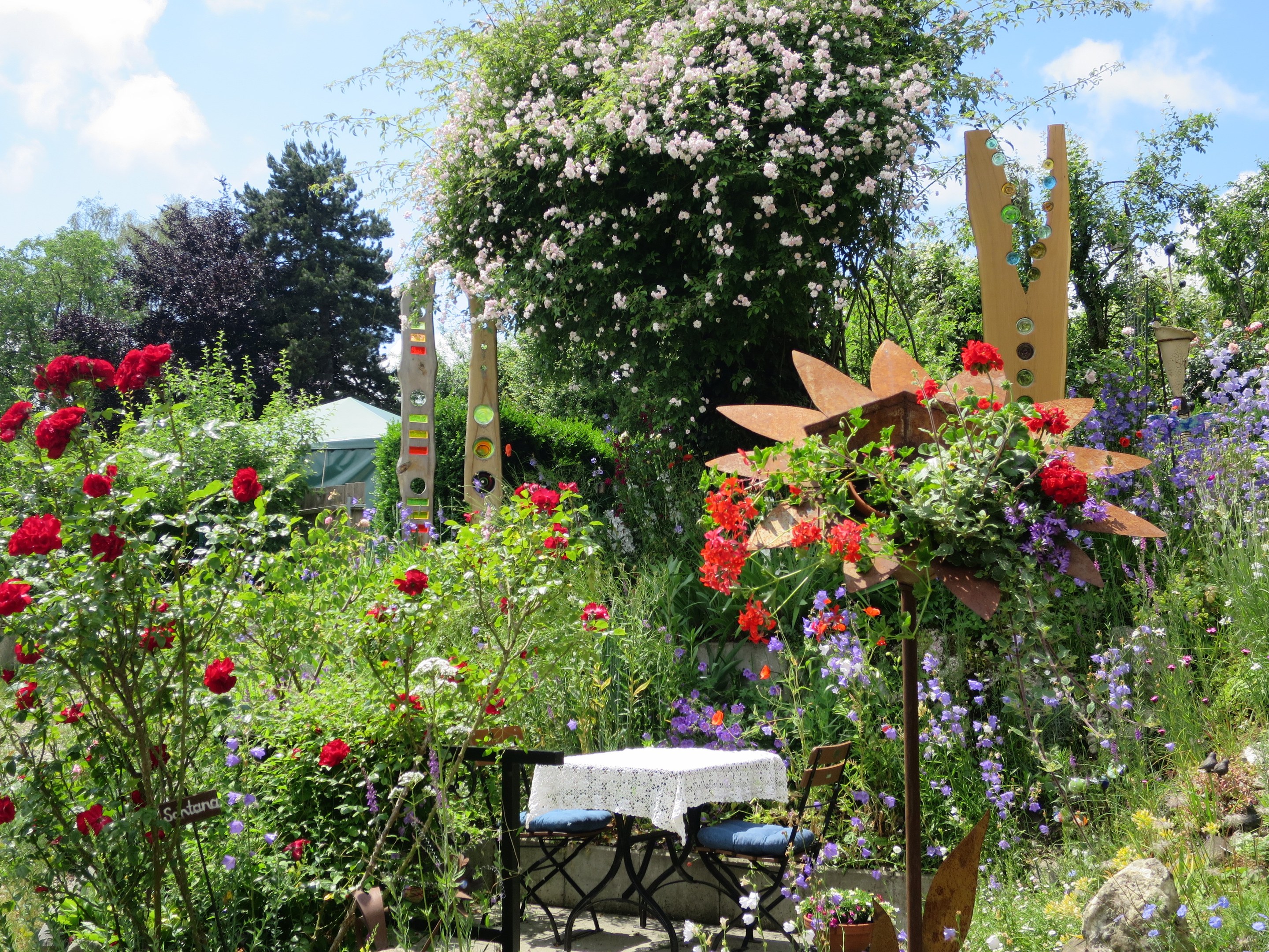 Ein Garten mit bunten Blumen und Pflanzen, der einen Tisch mit einer Decke und Stühlen in der Mitte und umgeben von Bäumen unter einem bewölkten Himmel auf der Chelsea Flower Show zeigt.