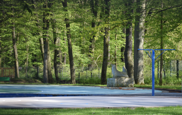 Basketballplatz in einem Park mit Bäumen, einer Bank links, einem Zaun im Hintergrund und Gras unten.