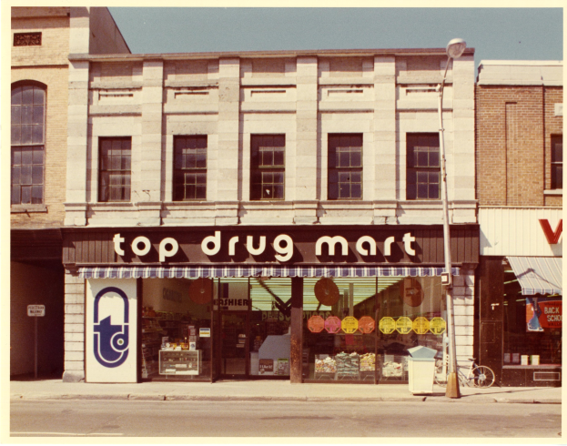 Ein Drogenmart-Laden an einer Straßenecke mit Fenstern, einem Schild, einem Straßenpfosten, einem geparkten Fahrrad, einem Müllcontainer und einem bewölkten Himmel.
