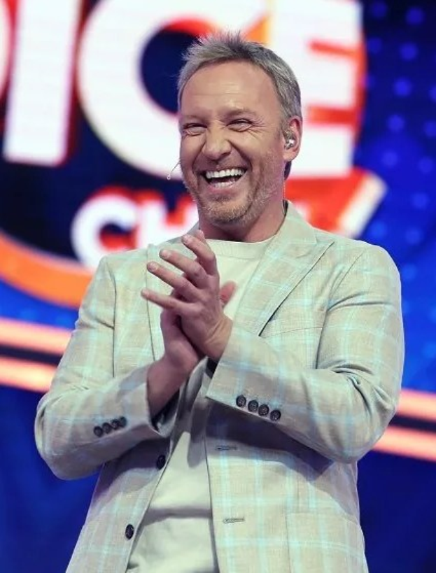 Ein Mann in einem Anzug klatscht auf einer Bühne mit dem 'The Voice'-Schild im Hintergrund.