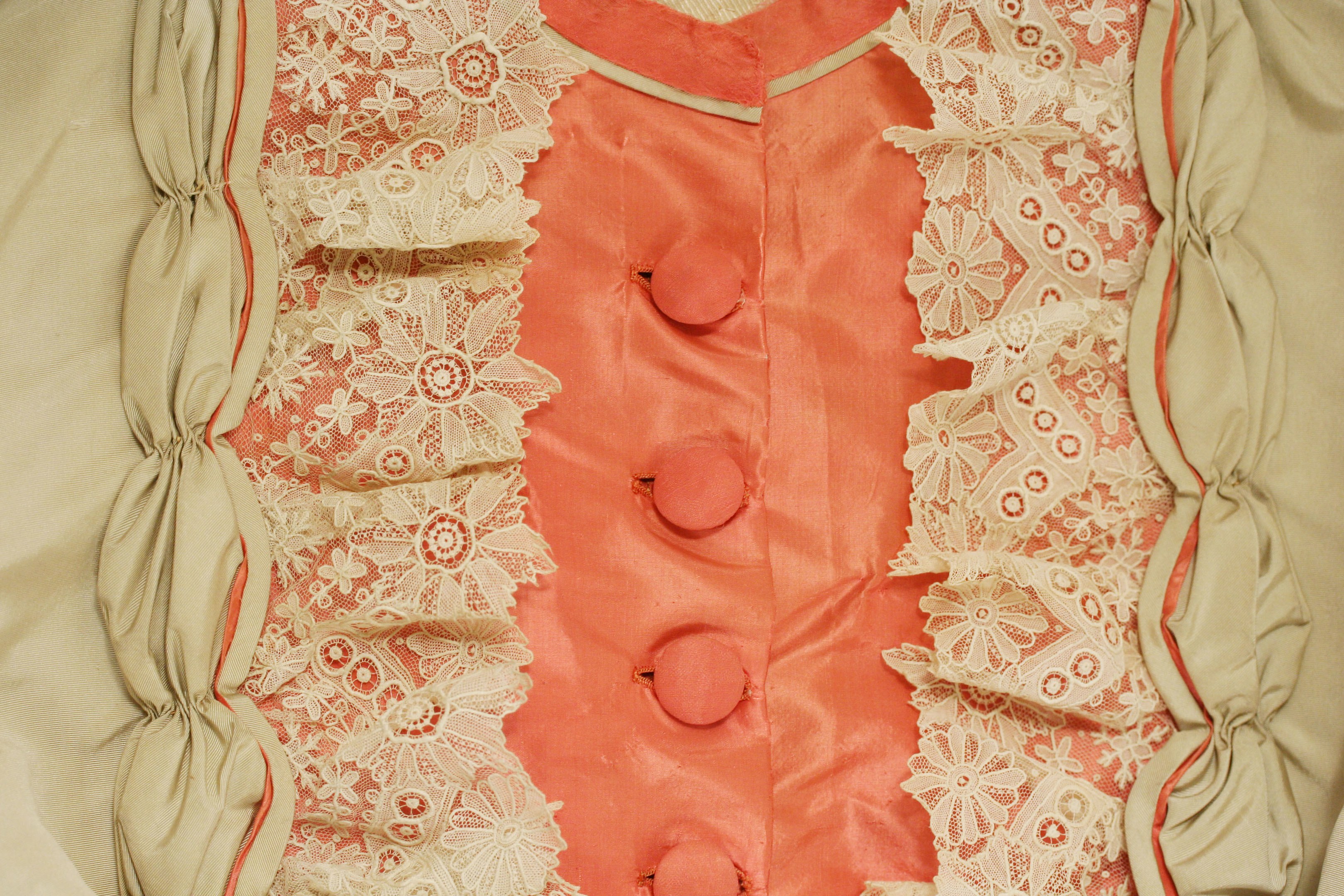 Nahaufnahme eines rosafarbenen und cremefarbenen Kleides mit weißer Spitze und Knöpfen vorne, das eine klassische und elegante Gestaltung aufweist.