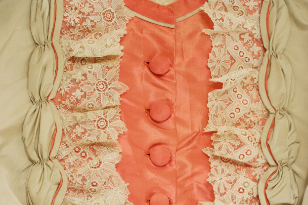 Nahaufnahme eines rosafarbenen und cremefarbenen Kleides mit weißer Spitze und Knöpfen vorne, das eine klassische und elegante Gestaltung aufweist.