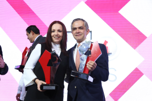 Zwei Personen auf einer Bühne, die lächeln und Trophäen halten, mit einem sichtbaren Bildschirm im Hintergrund.