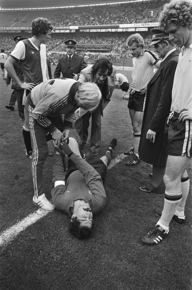 Schwarzes und weißes Bild eines Fußballspielers, der auf dem Boden liegt und von einer Gruppe von Männern umgeben ist, mit einer Menge im Hintergrund.