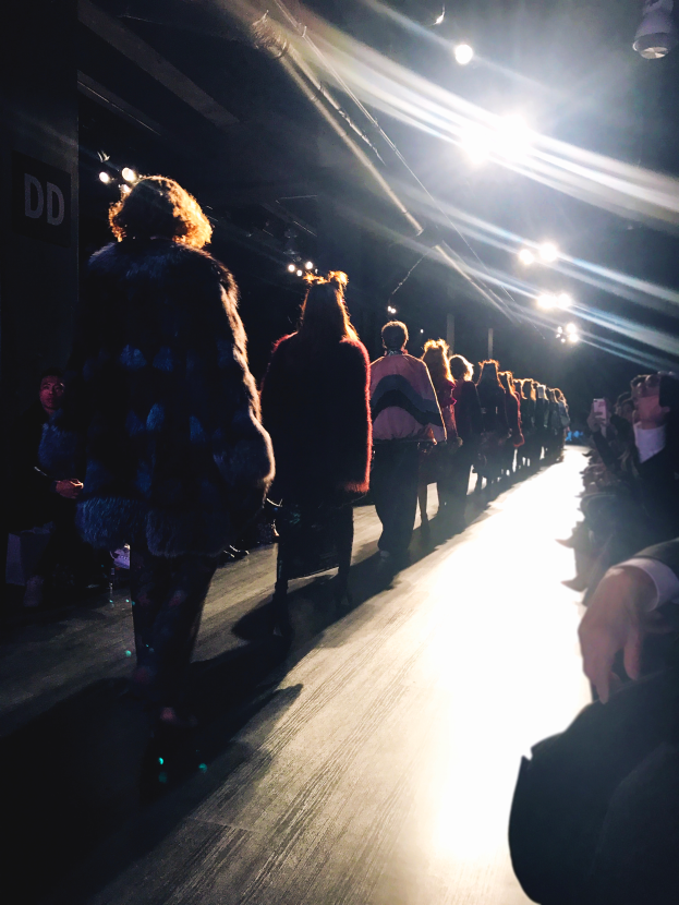 Eine Gruppe von Menschen läuft über einen Laufsteg bei einer Modenschau, beleuchtet von hellen Scheinwerfern, während Zuschauer auf beiden Seiten die Show beobachten.