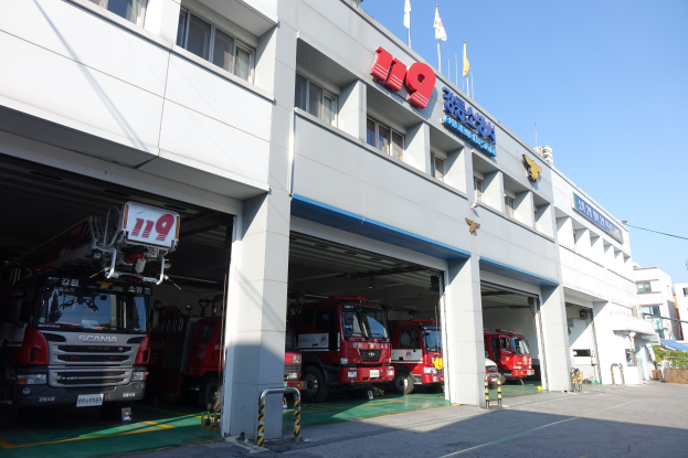 Feuerwache mit mehreren Feuerwehrautos vor dem Gebäude mit Fenstern, Säulen und Namensschildern, Flaggen im Vordergrund und anderen Gebäuden gegen einen klaren blauen Himmel.