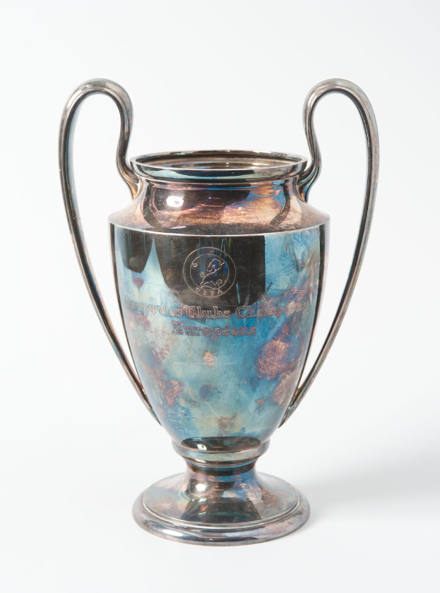 Der UEFA-Champions-League-Pokal, ein stählerner Becher mit zwei Griffen und einem blauen und silbernen Farbschema, der mit "UEFA Champions League" beschriftet ist und vor einem weißen Hintergrund steht.