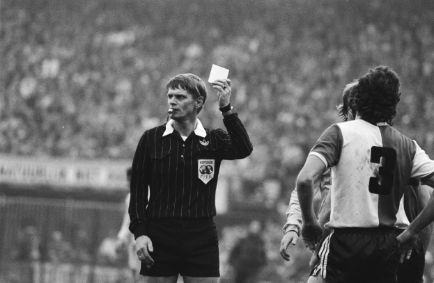 Schwarzes und weißes Bild eines Schiedsrichters mit roter Karte vor einer Menge in Fußballtrikots mit einem Zaun im Hintergrund.