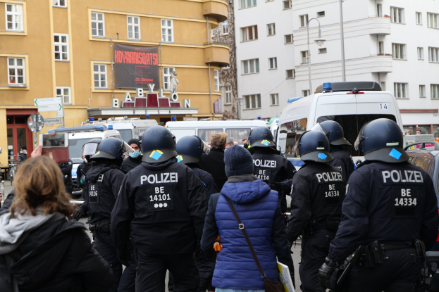 Polizisten in Uniform vor einer Menge bei einer Demonstration in Berlin, mit Fahrzeugen, Gebäuden und einem Banner im Hintergrund.