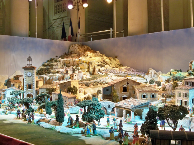 Ausgestelltes detailgetreues Modell einer Krippe in einem Museum mit Figuren, Gebäuden, Bäumen, Felsen, einem Glockenturm, Fahnen, Pfählen, Lichtern und einer Wand, beleuchtet vom Museumslicht.