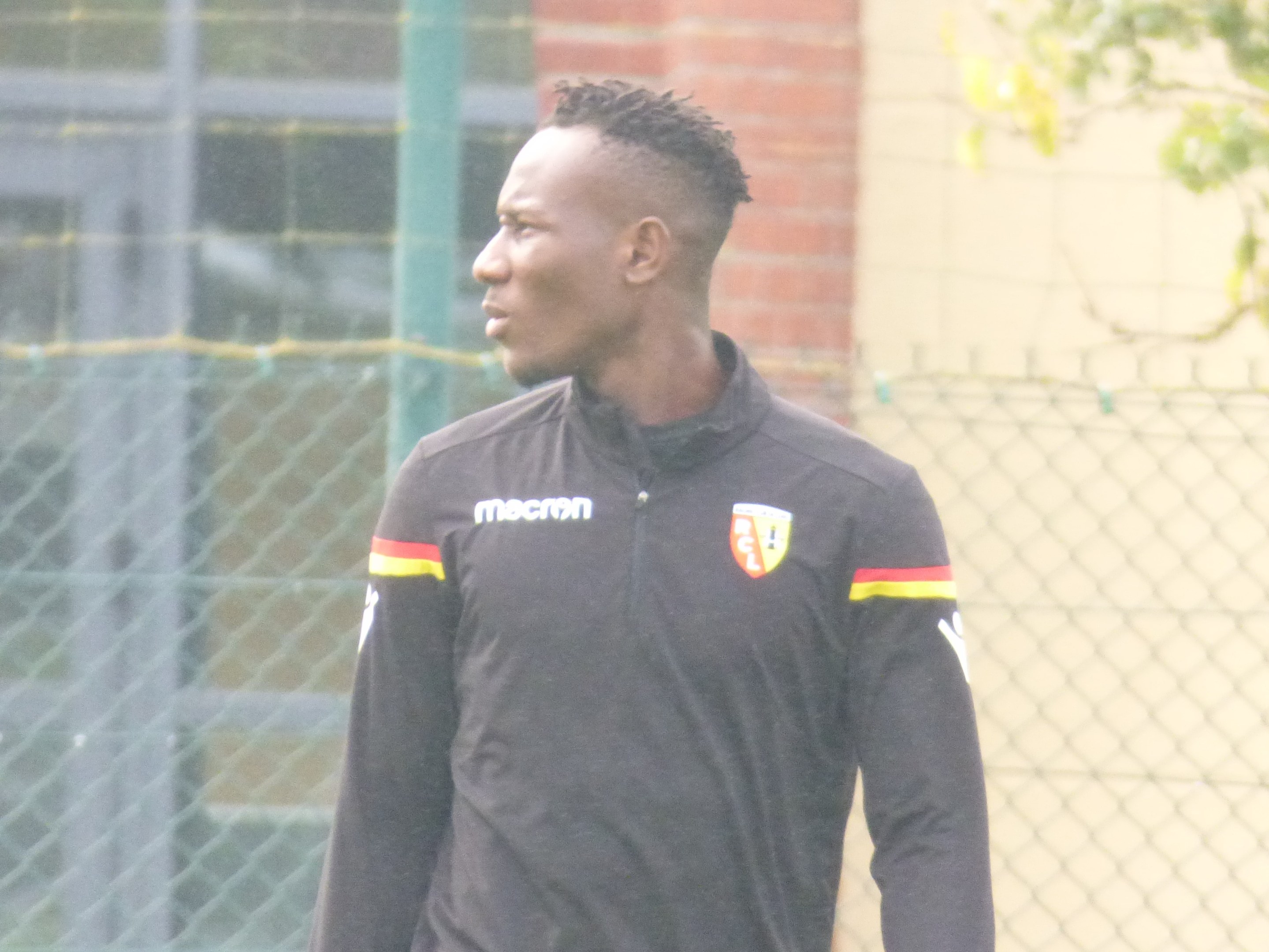 Ein Mann in einer schwarzen Jacke steht auf einem Fußballfeld, umgeben von einem Zaun und einem Baum, und sieht konzentriert während eines Trainings.