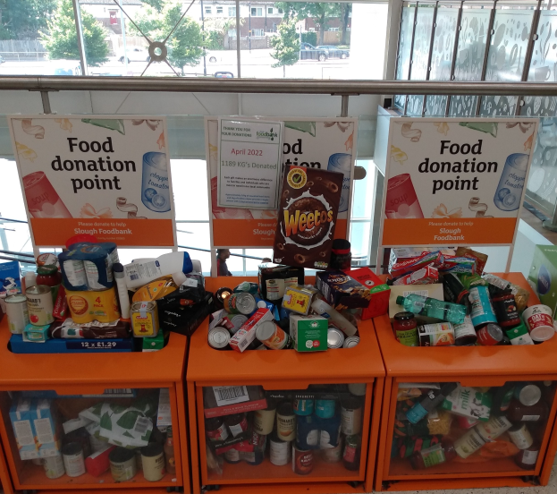 Orange Schränke mit Lebensmittelspenden gefüllt, darunter Flaschen, Dosen und Kartons, mit Informationsschildern und einem Blick durch Glasfenster auf Fahrzeuge, Bäume, Gebäude und Himmel.