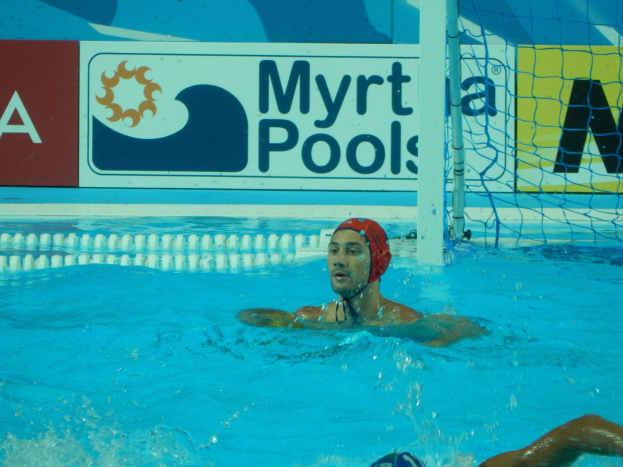 Wasserballspieler in Bewegung in einem Pool, trägt eine Mütze, mit einem Netz auf der rechten Seite und einem Werbeplakat im Hintergrund.