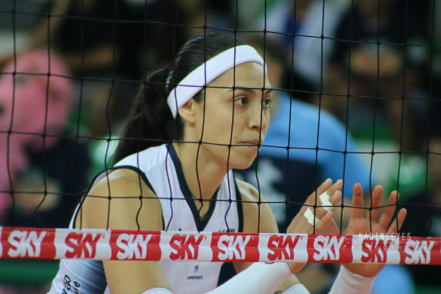Eine Volleyballspielerin in Uniform ist im Einsatz auf dem Feld, mit einem Netz im Vordergrund und einem unscharfen Hintergrund, der eine entschlossene Miene zeigt.