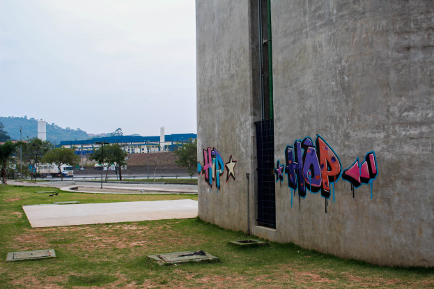 Betongebäude mit Graffiti an der Seite, vor einem Hintergrund aus Gras, Bäumen, Strommasten, Fahrzeugen auf der Straße, fernen Gebäuden, Hügeln und einem klaren blauen Himmel.