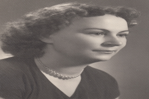 Ein Schwarz-Weiß-Foto einer Frau mit ernstem Gesichtsausdruck, die eine Kette und ein einfaches Kleid mit hohem Kragen und langen Ärmeln trägt, ihr Haar ist zurückgebunden und ihre Augen sind konzentriert.