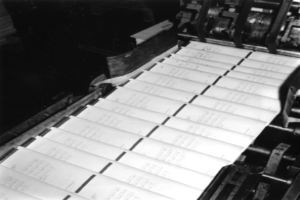 Ein Schwarz-Weiß-Foto von einer Druckmaschine mit mehreren Papierbögen darauf.