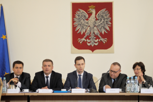 Eine Gruppe von Menschen sitzt an einem Tisch mit Namensschildern, Flaschen, Mikrofonen, Tassen und Untertassen, vor zwei Flaggen, mit einem gerahmten Objekt im Hintergrund, das wie ein Parlamentsbeamter aussieht.