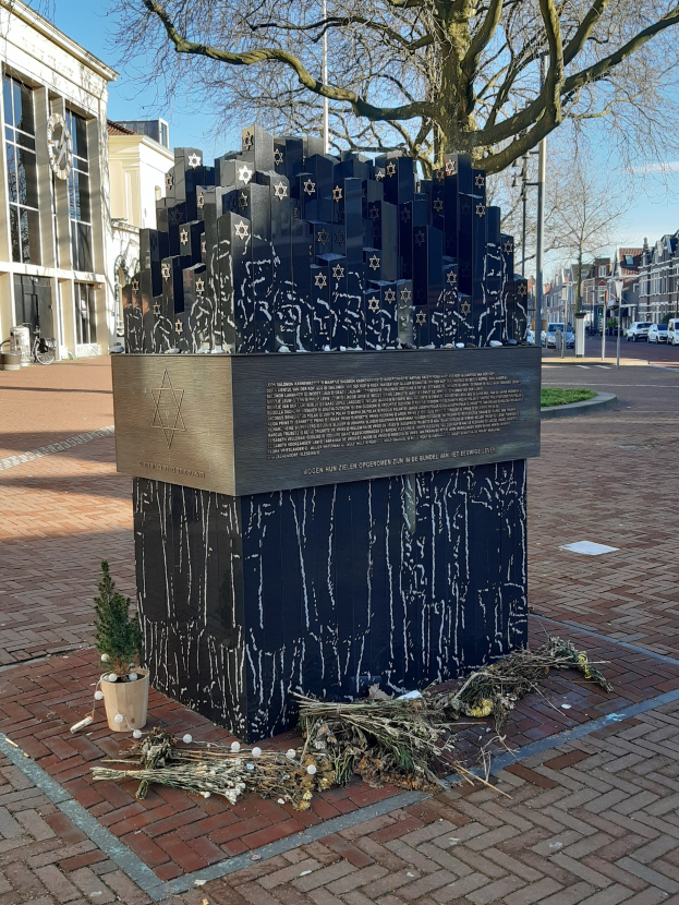 Denkmal für Holocaust-Opfer in einem Stadtplatz mit einem Baum, Topfpflanze, verstreuten trockenen Blättern und urbanem Hintergrund mit Gebäuden, Fahrzeugen, Masten und Bäumen.