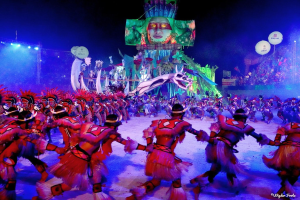 Eine Gruppe von Menschen in farbenfrohen Kostümen tanzt auf einer Bühne vor einem sitzenden Publikum, mit Lichtern, Pfählen und Ballons, und ein Bildschirm zeigt eine Person im Hintergrund, während des Karnevals in Rio de Janeiro 2014.