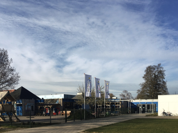 Gro├če Geb├Ąude mit Metallzaun, Rasenfl├Ąche, Weg, B├Ąume, Masten, Schilder, parkendes Fahrrad, n├Ąhere H├Ąuser und bew├Âlktem Himmel, identifiziert als neuer Spielbereich für Kinder an der Schule.