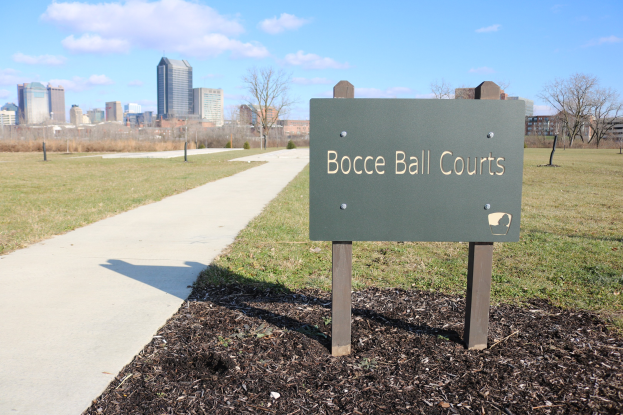 Ein Schild am Boden umgeben von Gras, einem Weg, Bäumen, Pfählen, einem Zaun, Gebäuden und einem bewölkten Himmel trägt die Aufschrift "Bocce Ball Courts in Indianapolis, Indiana".