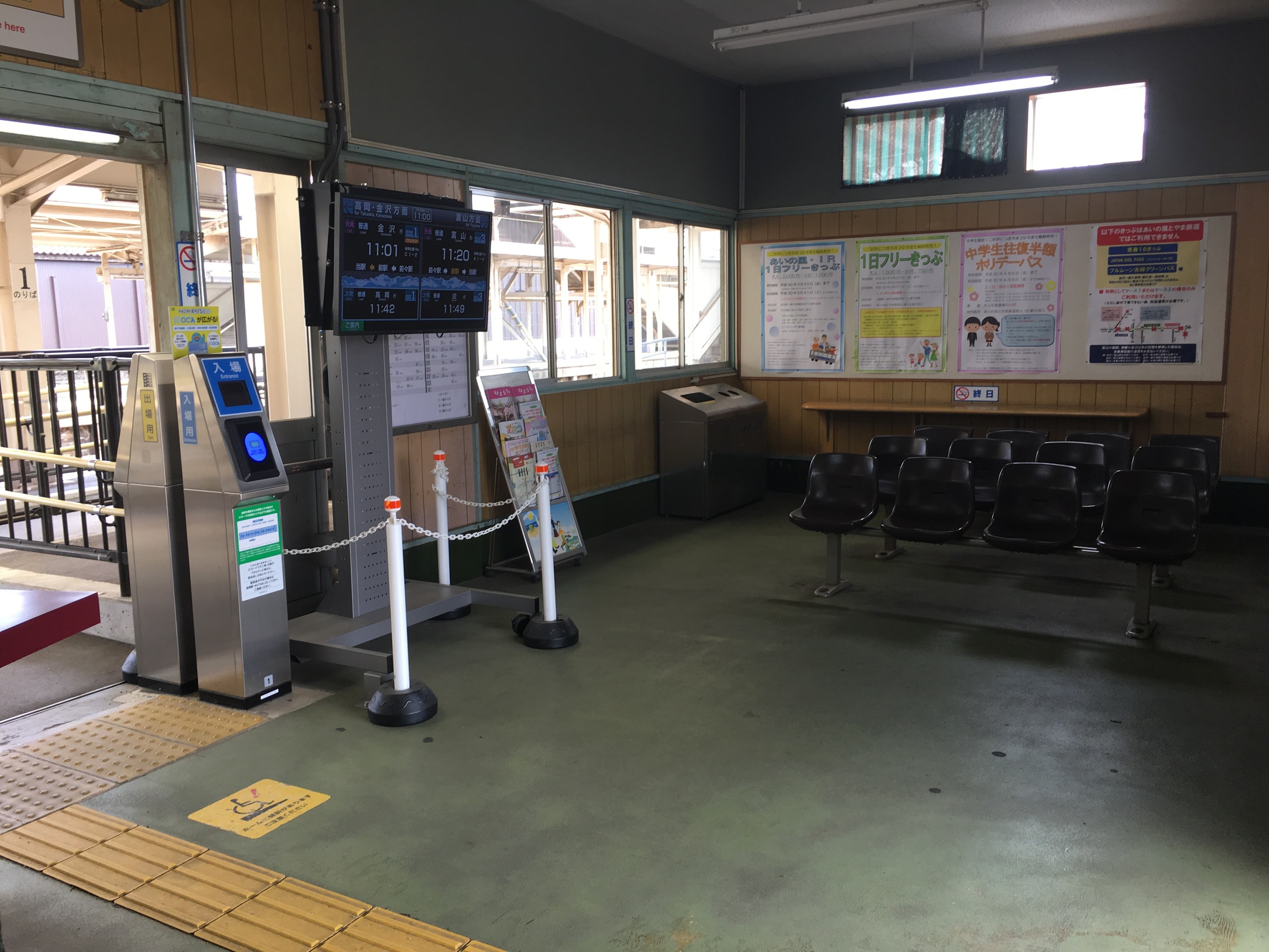 Ein belebter Bahnhofseingang mit Reihen von Stühlen, einem Service-Schalter, Ticketautomaten, Kettenbarrieren, Informationsanzeigen, Wandplakaten, Deckenbeleuchtung und Geländern.