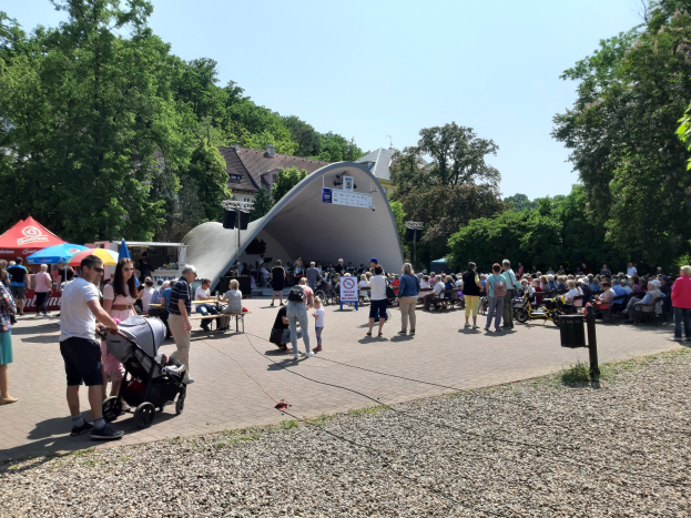 Eine Menschenmenge versammelt sich vor einem Gebäude bei der Festival-Eröffnung, einige sitzen auf Bänken, andere stehen; ein Kinderwagen ist im Vordergrund, und Bäume, Gebäude und ein klarer blauer Himmel sind im Hintergrund zu sehen. Zelte säumen die linke Seite, und ein Müllcontainer ist auf der rechten Seite zu sehen.