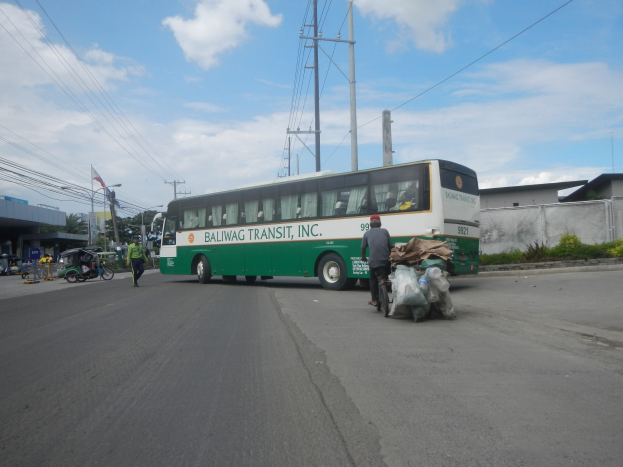 Ein grün-weißer Bus führt die Straße entlang neben einem Gebäude, mit Fußgängern und einem Radfahrer in der Nähe, vor dem Hintergrund von Strommasten, Gebäuden, Bäumen und einem bewölkten Himmel.