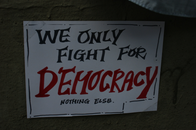 Weißes Papier mit "Wir kämpfen nur für Demokratie" darauf, an einer Wand befestigt.