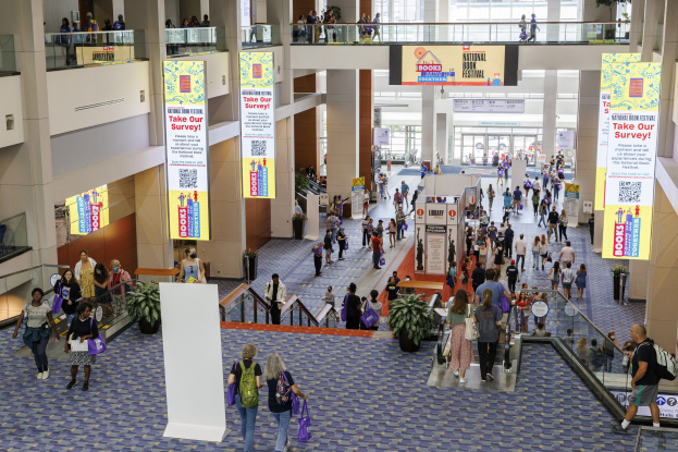 Eine große Menschenmenge geht durch ein großes Gebäude mit Bannern, Topfpflanzen, Rolltreppen, Geländern, Säulen, bedruckten Schildern und Glastüren, einige tragen Taschen, was auf den Besuch der Nationalen Buchmesse hinweist.
