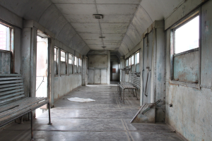 Altes Zuginnere mit Holzsitzen, Fenstern auf beiden Seiten und einer Decke, mit einem Gebäude im Hintergrund.