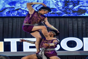 Zwei Performer in purpurfarbenen Kleidern führen Akrobatik auf der Bühne aus, während ein Mann eine Frau hochhebt und das Publikum darunter zusieht.