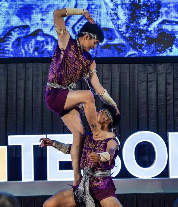 Zwei Performer in purpurfarbenen Kleidern führen Akrobatik auf der Bühne aus, während ein Mann eine Frau hochhebt und das Publikum darunter zusieht.