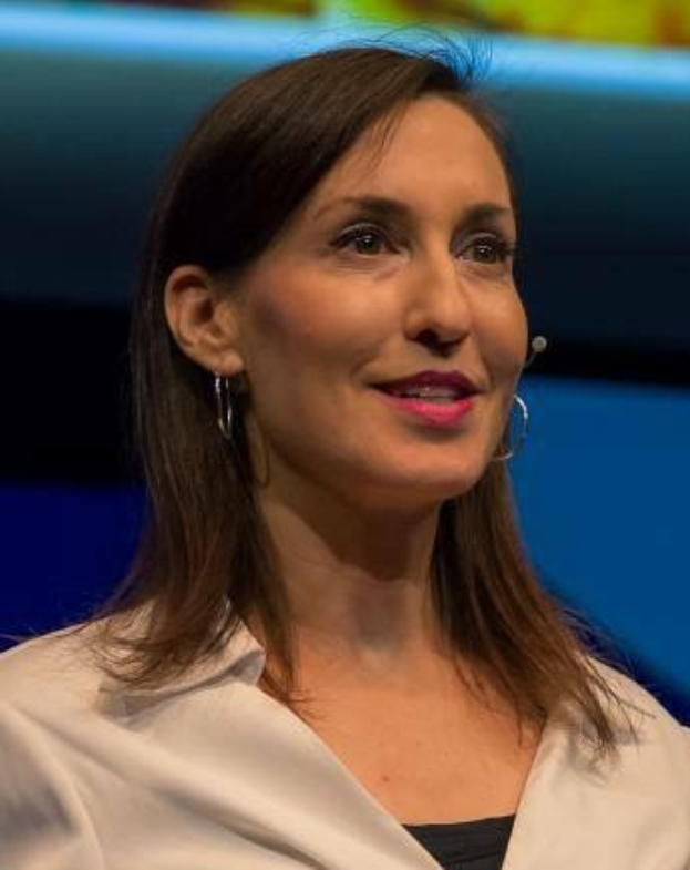 Frau in weißer Jacke und schwarzem Oberteil vor einem blauen und gelben Hintergrund sitzend, mit Ohrringen und einem Mikrofon während eines Interviews.