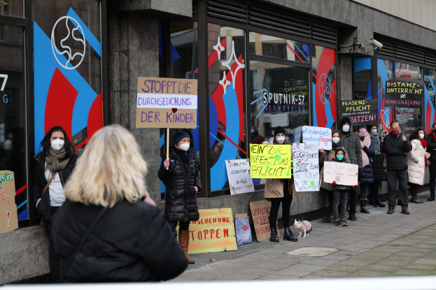 Gruppe von maskierten Menschen mit Schildern und Plakaten vor einem gläsernen Gebäude, mit Kameras und einem Hund, wahrscheinlich protestierend gegen eine deutsche Regierung Entscheidung, Masken in Schulen zu verbieten.