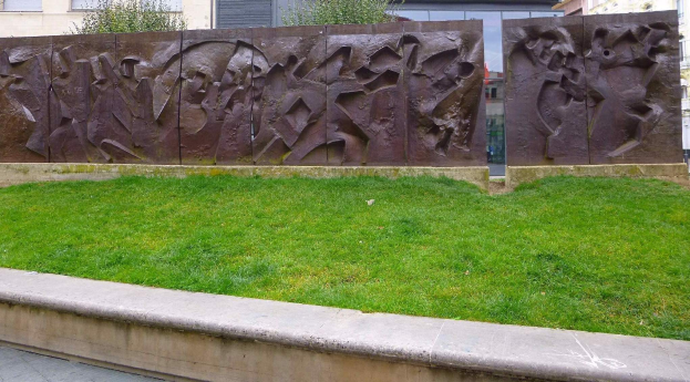 Große Metallskulptur der Berliner Mauer auf einer grünen Fläche umgeben von Bäumen und Gebäuden mit Fenstern im Hintergrund.
