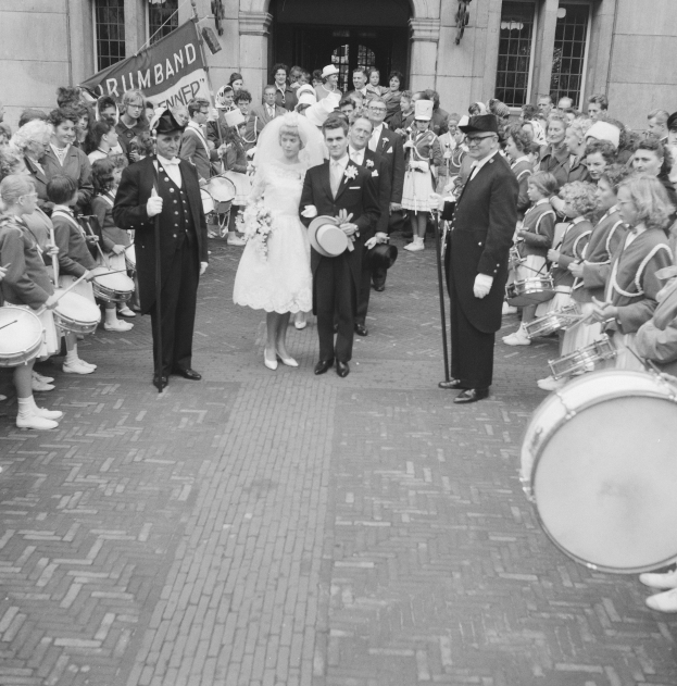 Schwarz-weißes Foto einer Braut in einem weißen Kleid und Schleier und eines Bräutigams mit einem Blumenstrauß, der eine Straße entlang geht, gefolgt von einer Menge mit Musikinstrumenten, vor einem Gebäude mit einer Fahne.