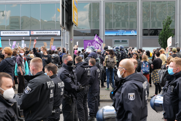 Große Gruppe von Menschen protestiert vor einem Gebäude, einige halten Schilder und tragen Helme, mit einem Schildständer und einem Baum im Hintergrund.
