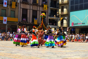 Eine Gruppe Frauen in bunten Kleidern tanzt auf der Straße, umgeben von einer Menschenmenge, mit Gebäuden, Geländern, Balkonen, Fahnen, einem Laternenmast und einer Tafel mit Schrift im Hintergrund.