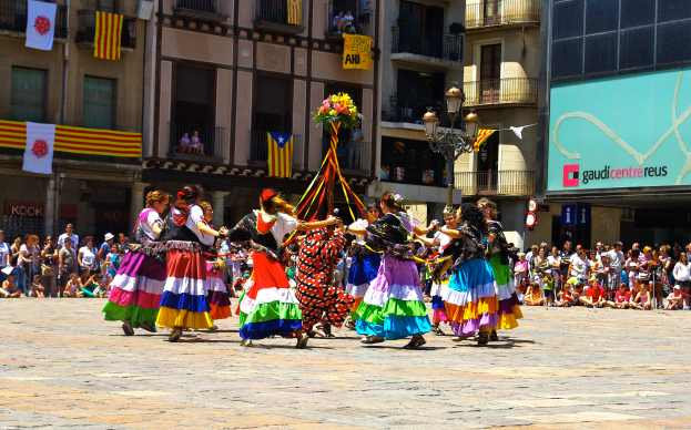 Eine Gruppe Frauen in bunten Kleidern tanzt auf der Straße, umgeben von einer Menschenmenge, mit Gebäuden, Geländern, Balkonen, Fahnen, einem Laternenmast und einer Tafel mit Schrift im Hintergrund.