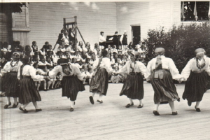 Eine Gruppe von Frauen tanzt vor einer Menge, mit einem Gebäude, Pflanzen und einem hölzernen Objekt im Hintergrund, alles in Schwarz-Weiß dargestellt.