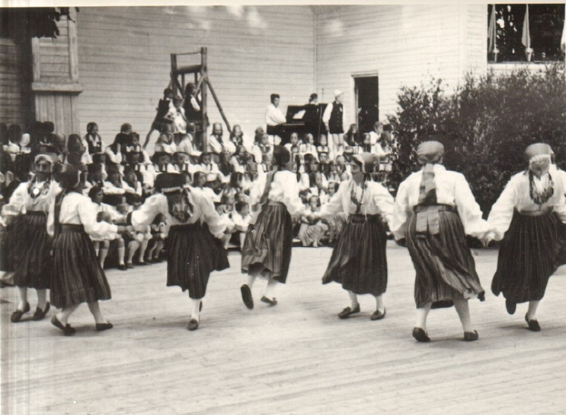 Eine Gruppe von Frauen tanzt vor einer Menge, mit einem Gebäude, Pflanzen und einem hölzernen Objekt im Hintergrund, alles in Schwarz-Weiß dargestellt.