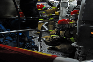 Feuerwehrleute in Schutzausrüstung arbeiten an einem Löschfahrzeug mit Schläuchen und einer Lampe im Hintergrund.