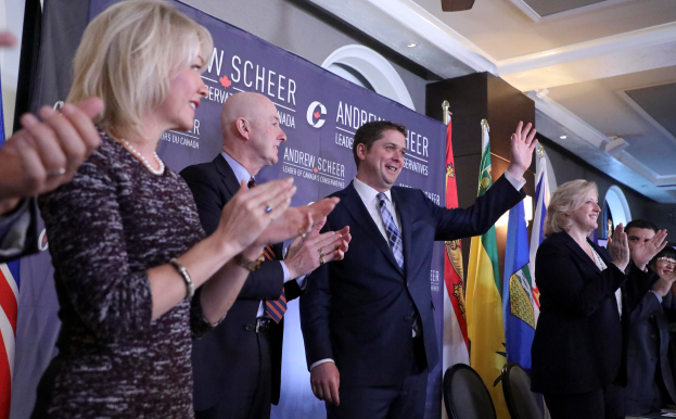 Eine Gruppe von Menschen, die vor Fahnen stehen und jubeln, mit einem Tisch voller Gegenstände rechts daneben, Stühlen, einer Fahne und einer Wand im Hintergrund und Lampen und einem Ventilator auf dem Dach.