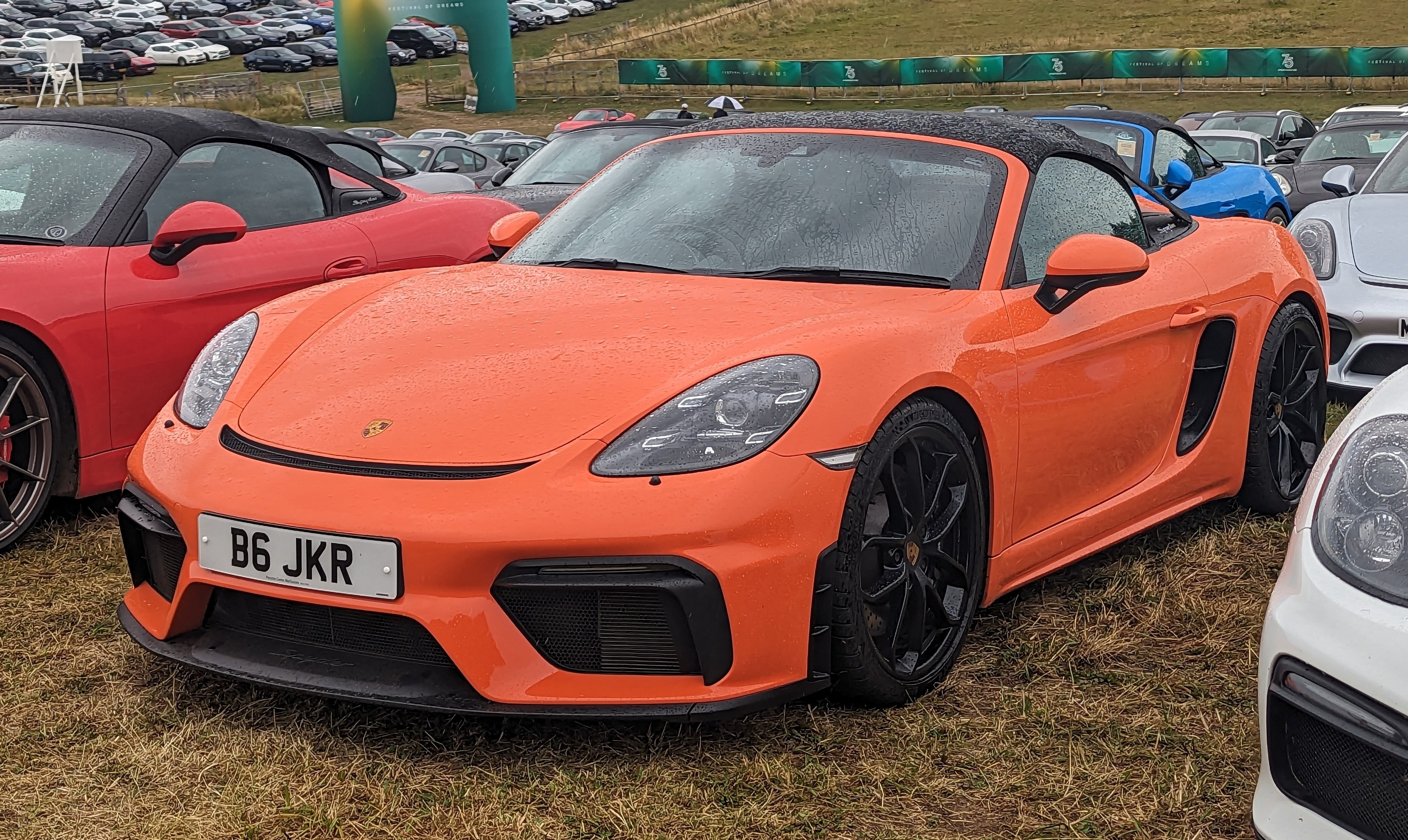 Roter Porsche 718 Boxster GTS auf der Goodwood Festival of Speed mit anderen Autos, einem Banner, Bäumen und einem klaren blauen Himmel im Hintergrund.