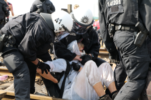 Eine Gruppe von Polizisten in schwarzer Einsatzkleidung verhaftet einen Mann in einem weißen Kleid auf einem Bahngleis, mit anderen Menschen im Hintergrund.